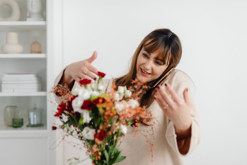 A woman with light brown hair and bangs, smiling warmly, is holding a vibrant floral bouquet that includes red, white, and orange flowers with green foliage. She is wearing a cream-colored sweater and