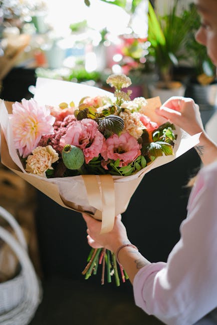 A person holding a large bouquet of fresh flowers wrapped in cream-colored paper, featuring pink peonies, white and purple chrysanthemums, and green foliage. The bouquet is arranged with a variety of
