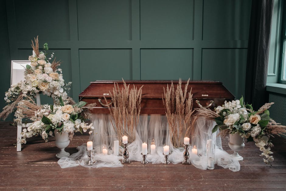A floral arrangement for a funeral, featuring large white roses, cream-colored carnations, and white lilies complemented by beige pampas grass, all arranged in white urn-style vases on either side of 