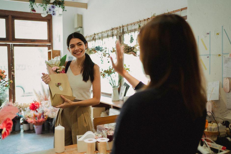 In a brightly lit flower shop, a young woman with dark hair tied back smiles warmly as she receives a floral gift from another person. She holds a bouquet encased in a light brown paper wrapping, feat