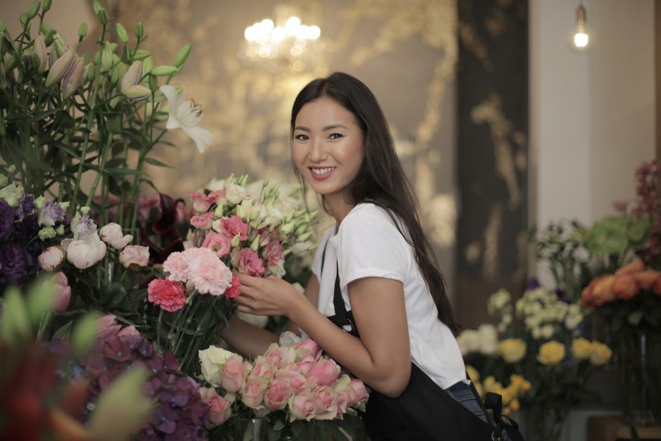 A young woman with long dark hair and a friendly smile stands amidst an arrangement of fresh flowers inside a florist shop. She is holding a bunch of pink and white carnations and is wearing a white T