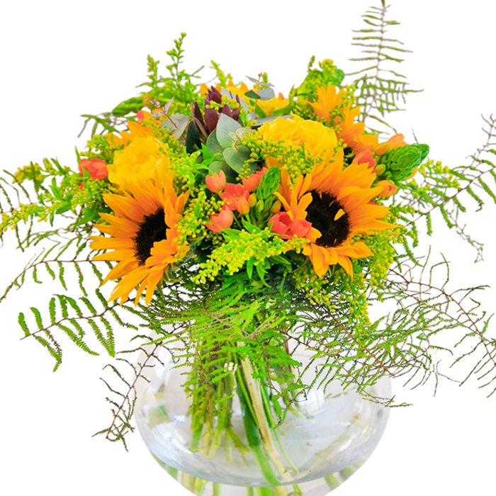 Exhibiting the abundant energy of a late summer meadow just outside Stevenage, this lush bouquet gathers radiant sunflowers and golden yellow roses in a clear, rounded glass vase. The sunflowers' broad, dark centres are framed by luminous yellow-orange rays, their heads turned as if to greet the daylight pouring into a Stevenage kitchen on a bright morning. Nestled beside them, elegant garden roses coil their petals in buttery-yellow spirals, offering subtle hints of their delicate scent. Between these bold blooms, glossy red hypericum berries catch the eye, their vibrant colour amplifying the warmth of the arrangement. Fern fronds reach gracefully outward, evoking the wild greenery of Aston or the leafy lanes near Benington, while plumes of goldenrod add gentle motion and flecks of soft yellow-green. Eucalyptus leaves, tinged with silvery blue, contribute a touch of rustic calm and fresh aroma, rounding out the textural harmony. The vase's round clarity showcases the overlapping stems and glimmers with water beneath-a quiet detail that suggests freshness and attentive florist care. Every flower, berry, and leaf seems thoughtfully placed to evoke the gentle brightness of Stevenage's summer landscapes, making this arrangement an ideal centrepiece for joyful gatherings, heartfelt congratulations, or simply brightening a loved one's day wherever they may be in the SG1 area.